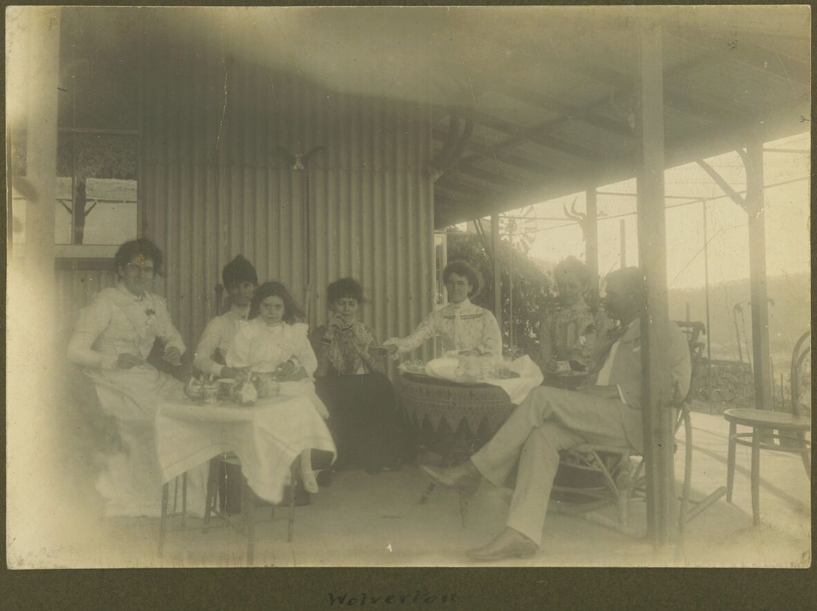 Faded vintage photo of a group having tea on a porch, dressed in early 1900s clothing.