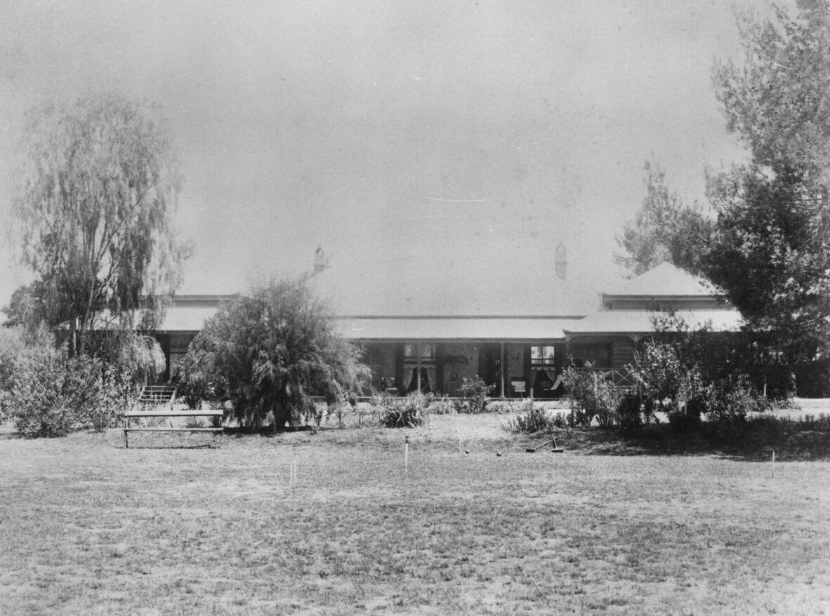 Black-and-white photo of a large house with a wide porch, surrounded by trees, shrubs, and an open grassy yard.
