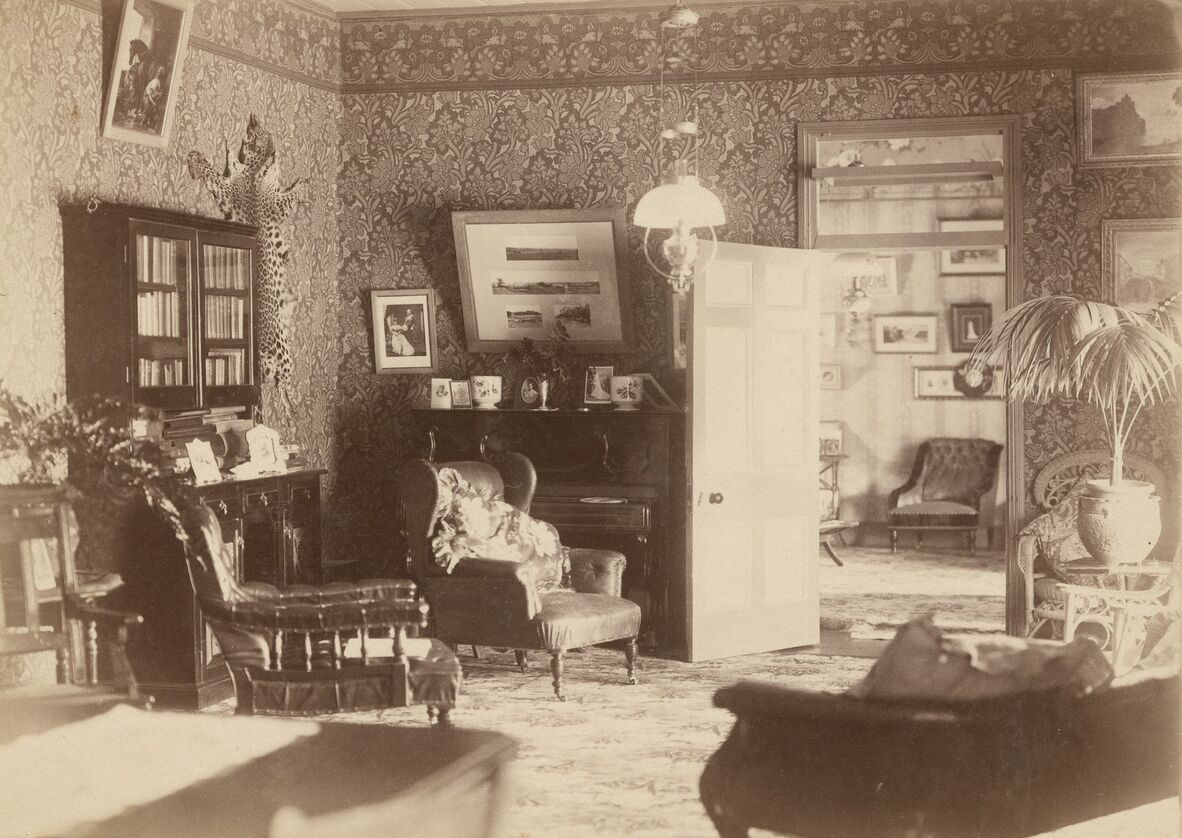 A sepia-toned photograph of a well-furnished early 20th-century living room featuring patterned wallpaper, upholstered armchairs, a piano with framed photographs on top, a glass-front bookcase, and decorative items including a mounted animal skin.