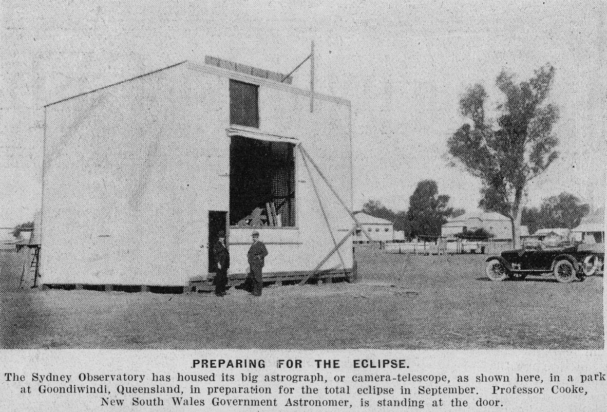A large box-like structure stands in a park, with two men beside it. An old car is parked nearby. The sky is clear, suggesting a daytime setting. The photo caption reads: "PREPARING FOR THE ECLIPSE. The Sydney Observatory has housed its big astrograph, or camera-telescope, as shown here, in a park at Goondiwindi, Queensland, in preparation for the total eclipse in September. Professor Cooke, New South Wales Government Astronomer, is standing at the door."