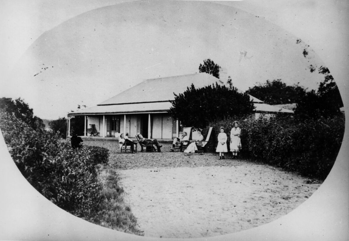 Black-and-white photo of a rural house with a porch, people relaxing in chairs, and two children standing nearby in a garden.