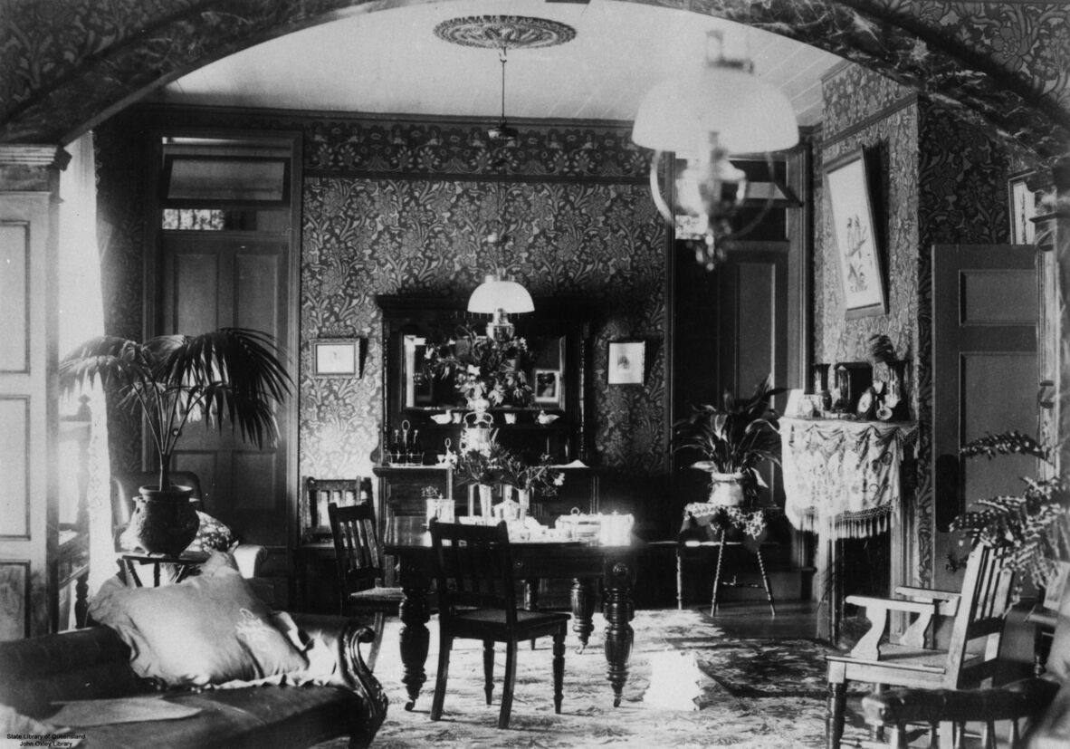 Black-and-white photo of a richly decorated living room with patterned wallpaper, wooden furniture, plants, and a central table under hanging lamps.