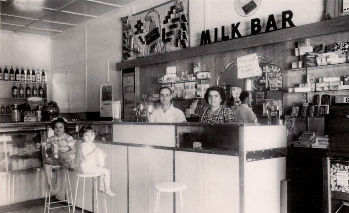 a man and woman behind the counter at a milk bar. two children sit on stools at the counter