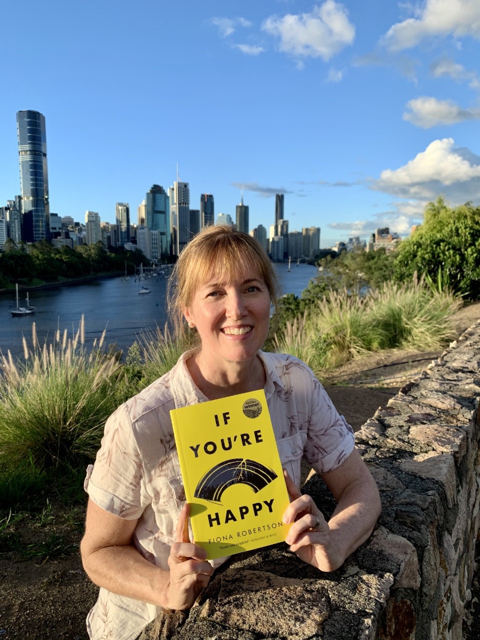 Fiona Robertson stands at Kangaroo Point cliffs holding a copy of her book. The river and city of Brisbane are behind her.