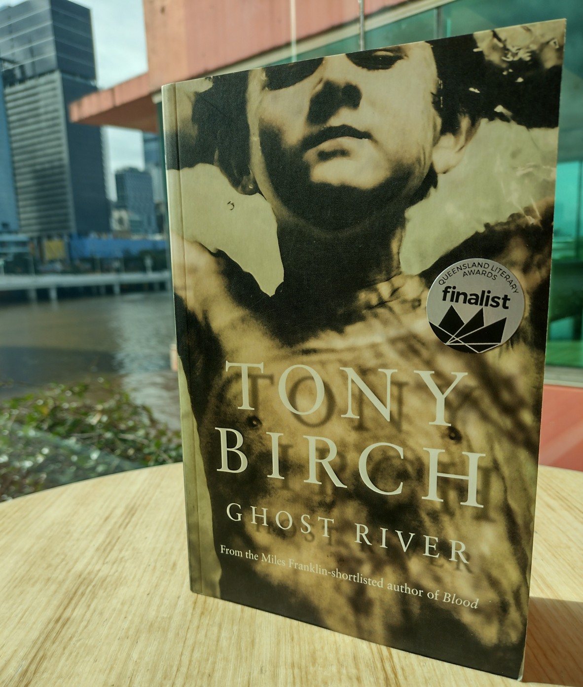 A book sitting on a table in a light-filled space with a cityscape and glimpse of a river in the background. The text on the cover reads Tony Birch Ghost River. From the Miles Franklin-shortlisted author of Blood.