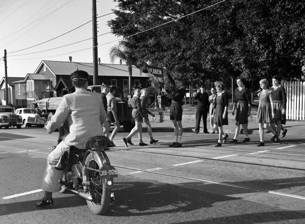 A police officer is standing to the side of the road and a student in the middle of the road is holding a Children Crossing sign as other schoolchildren cross. The church in the background is St Andrew's Anglican Church, Lutwyche. The church is located next to Woolowin State School.