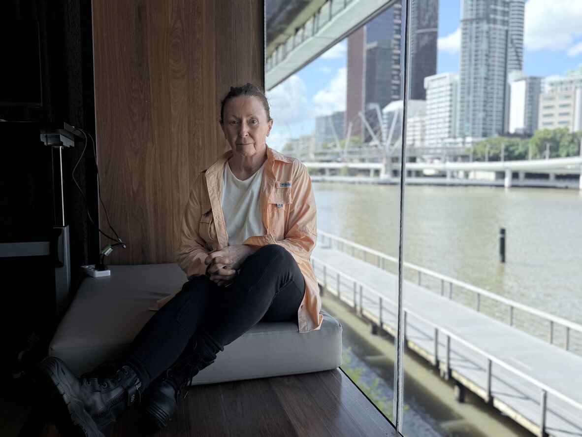 A woman in black jeans and an orange overshirt sits next to a window with the Brisbane river outside. She looks straight down the lens at the photographer