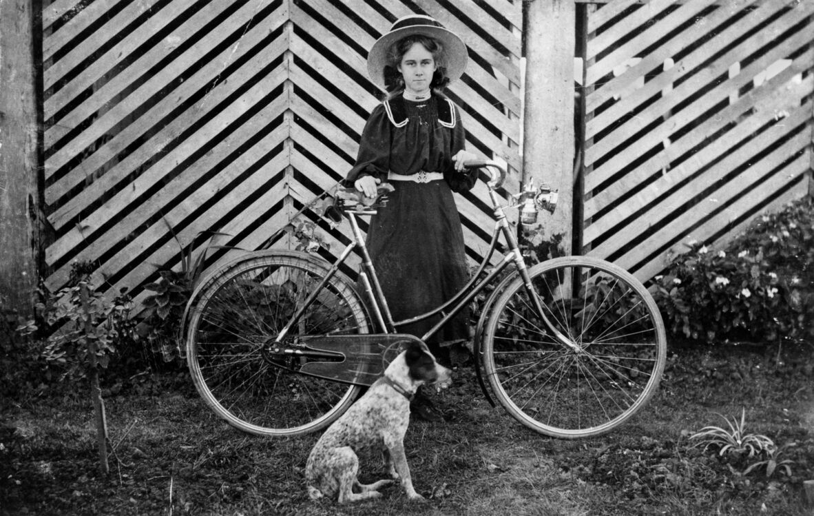 Black-and-white photograph of a young woman in early 20th-century dress and wide-brimmed hat standing beside a bicycle, holding the handlebars. A small dog sits at her feet. She poses in front of a patterned wooden fence with garden plants surrounding her.