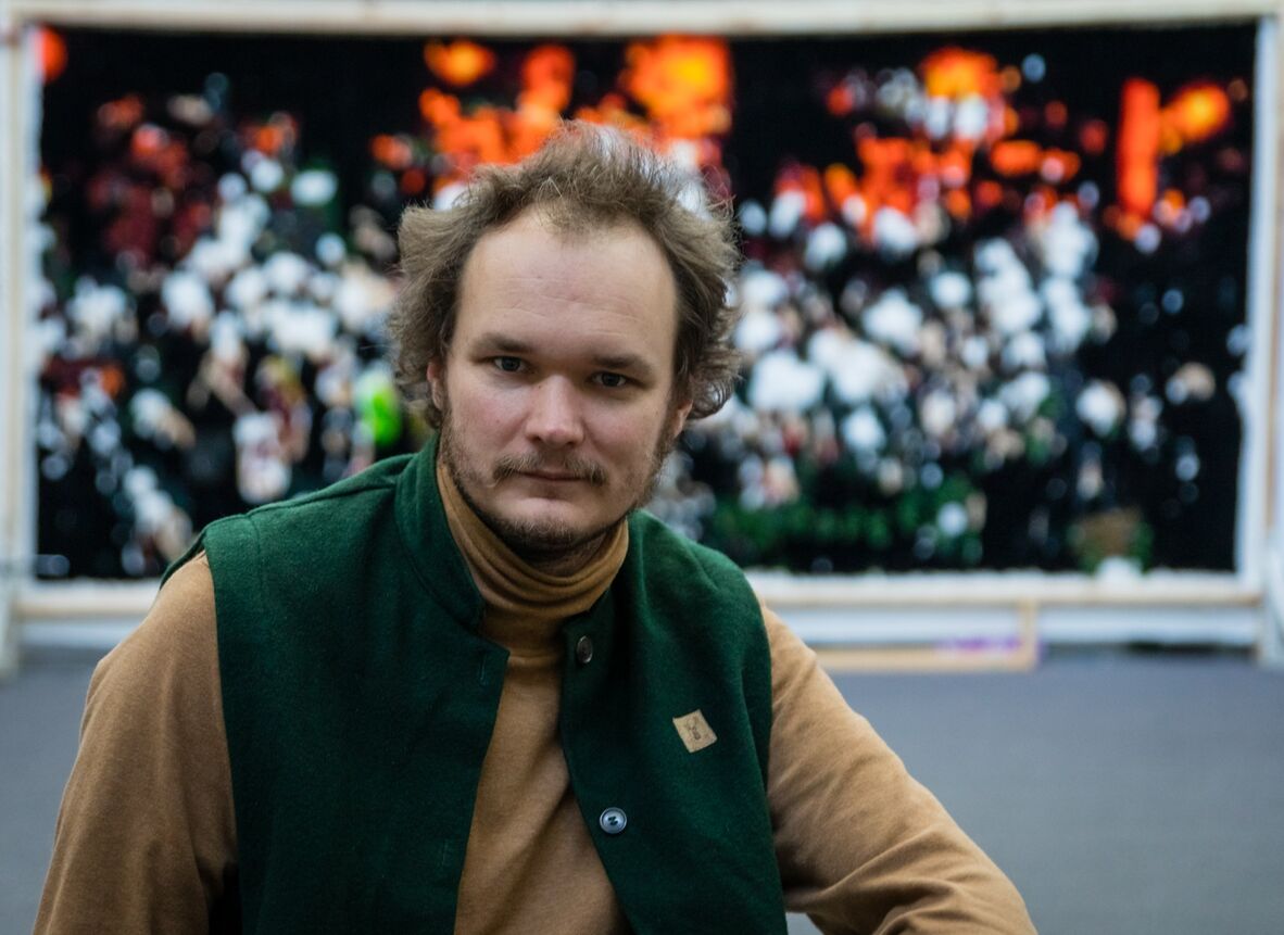 Joseph stands in front of a black background with blurry red and white lights in visible. Jo is on the left side of the foreground looking at the camera. He is wearing a light brown turtleneck and green vest.