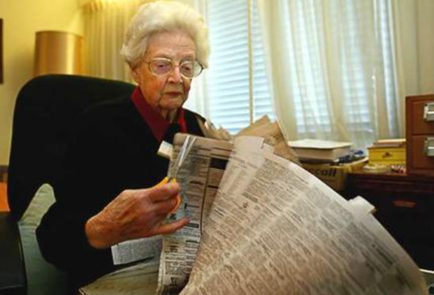 An older woman wearing glasses sitting reading a newspaper
