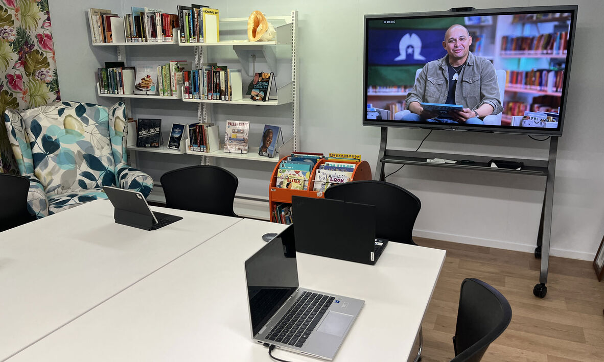 Laptops on a table with a large portable screen and book shelves in the background. 