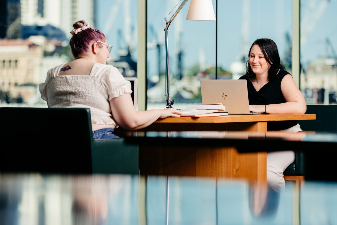 Two women sitting at a joint desk with a view of Brisbane city using laptops at State Library.