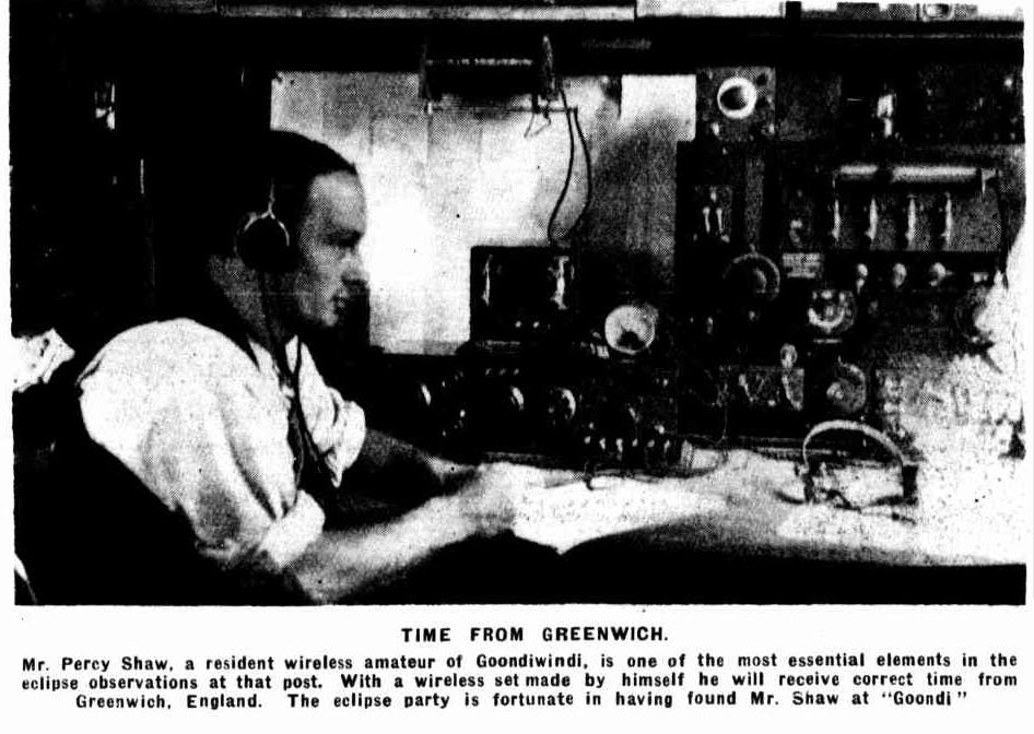 A man, wearing headphones, focuses intently on operating vintage radio equipment. Text reads: "TIME FROM GREENWICH. Mr. Percy Shaw, a resident wireless amateur of Goondiwindi, is one of the most essential elements in the eclipse observations at that post. With a wireless set made by himself he will receive correct time from Greenwich, England. The eclipse party is fortunate in having found Mr. Shaw at "Goondi"."