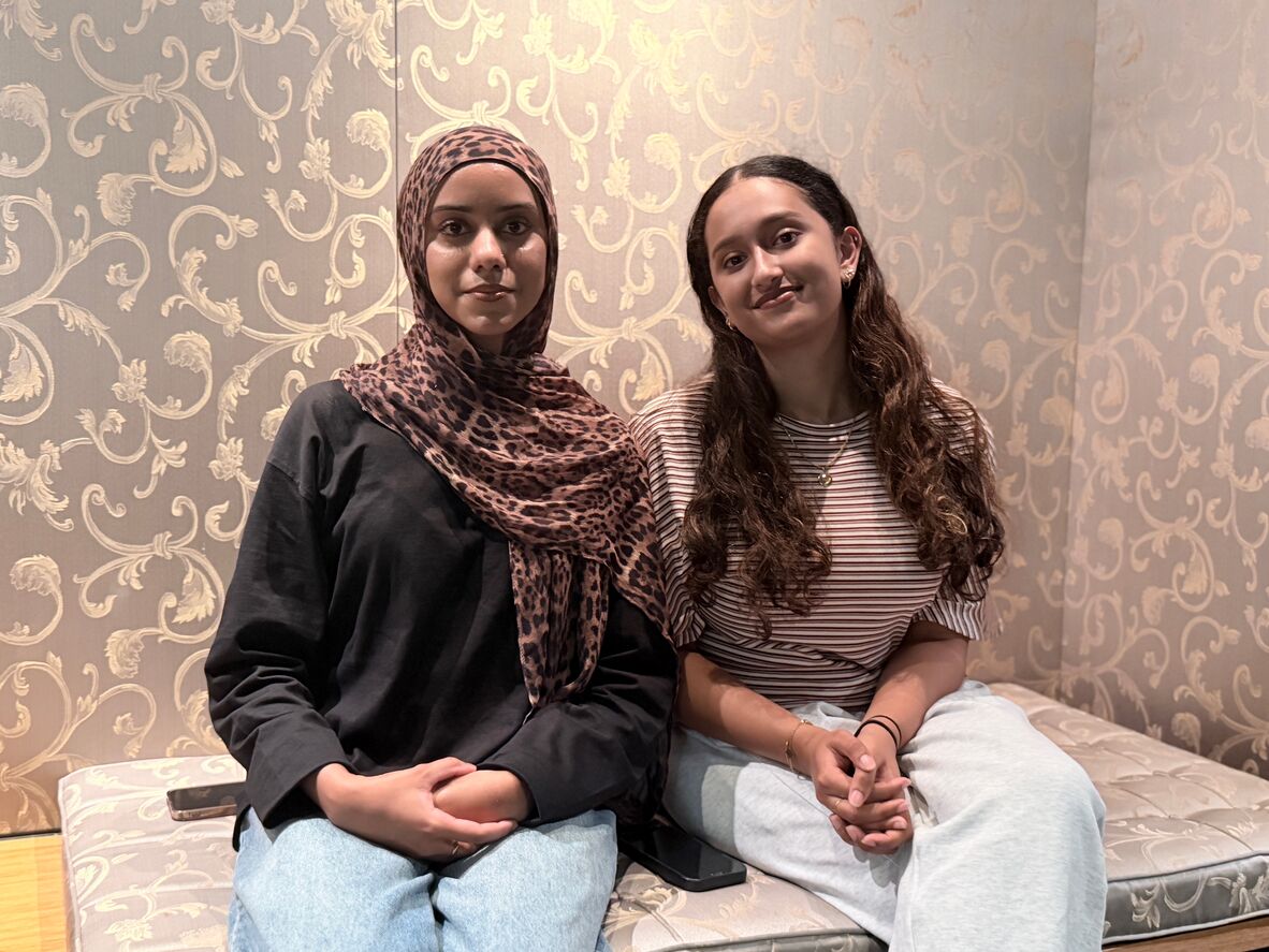 Two people sitting on a soft bench in front of a floral gold wallpaper. They are looking at the Camera, the one on the left is wearing a leopard print headscarf and the other has long curly brown hair and is simling