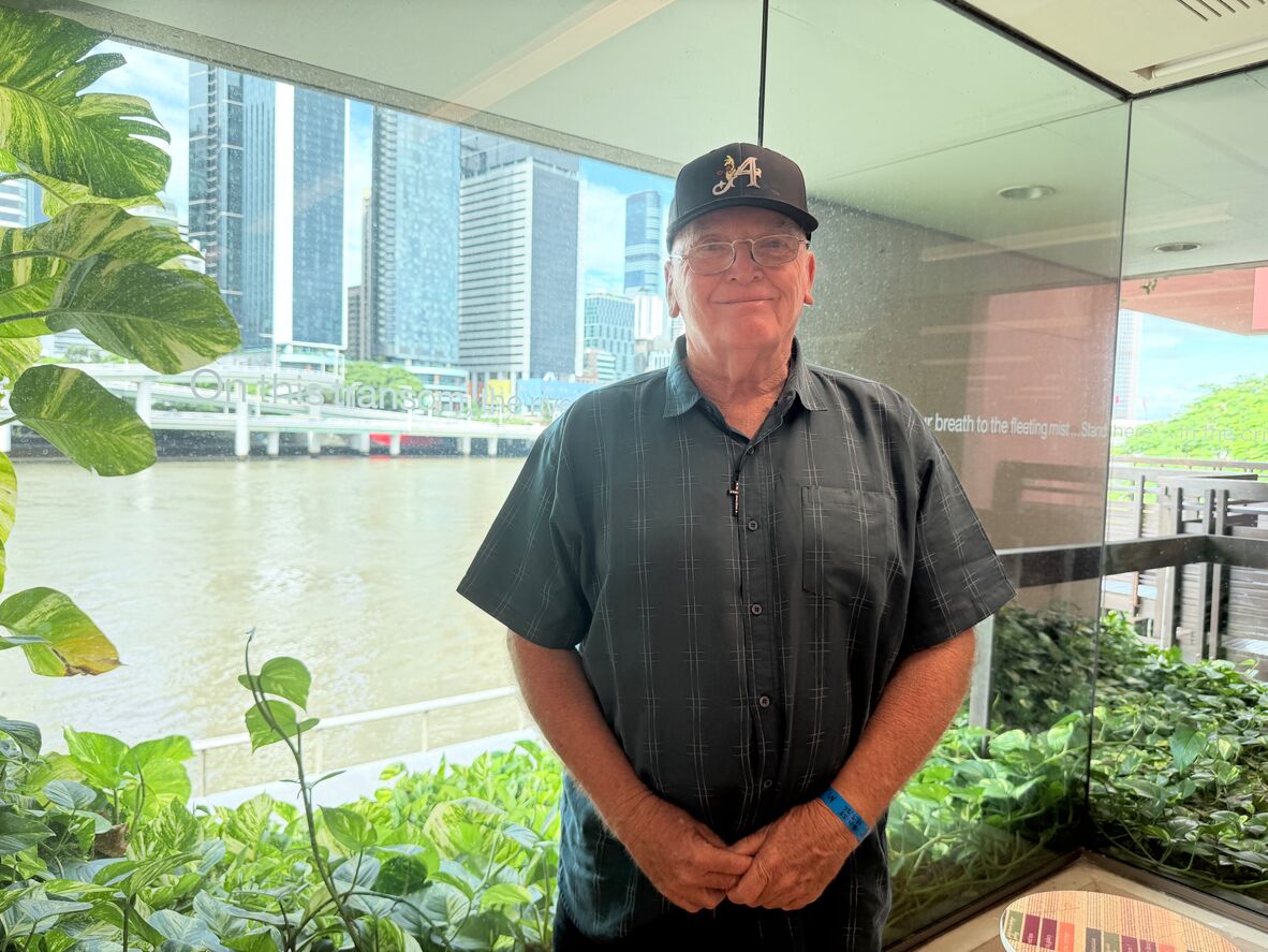 Greg stands in front of a window overlooking the Brisbane river, green plants can be seen all around him, he is wearing a button up, glasses and a black cap, he is looking at the camera and similing slightly.