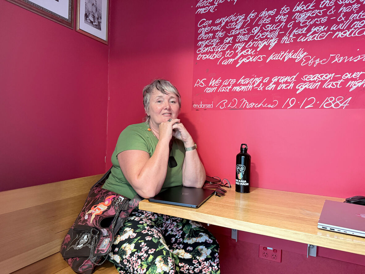 Ruth sits in the Black Opium reading room on Level 4, she is sitting in front of a bright pink wall leaning against the table built into the wall. Her arms are under her chin and her laptop is on the table below her, she is looking at the camera and wearing a green and floral outfit.