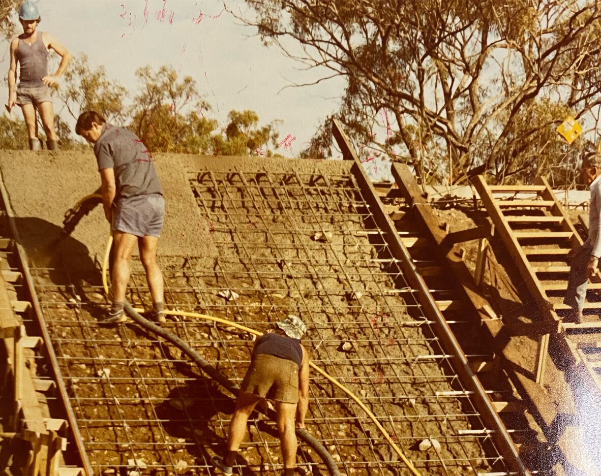 Colour photograph of workers constructing a velodrome, standing on a steep embankment lined with steel reinforcing mesh. One man operates a hose pouring concrete while others guide it. Timber formwork frames the slope, with trees and dry landscape visible behind the active construction site.