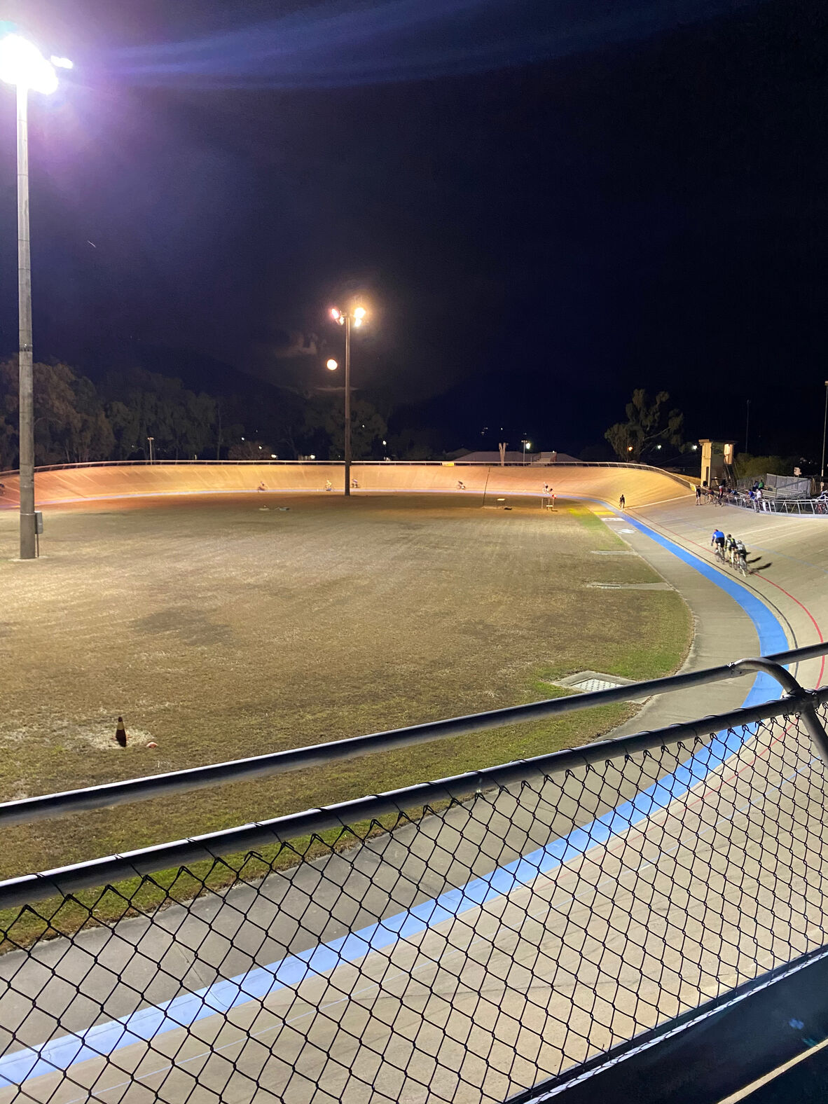 Night-time view of a velodrome illuminated by tall floodlights, with cyclists riding along the steeply banked track. A chain-link fence lines the foreground. Spectators gather near the track edge, while the grassy infield sits dark beneath a cloudy sky and visible moon.