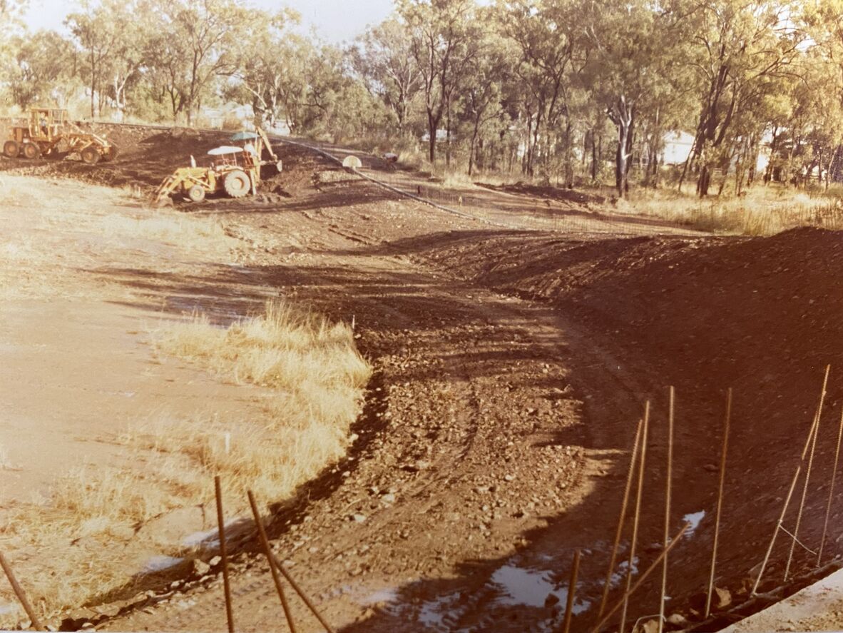 Faded colour photograph showing construction of a velodrome track, with earthmoving machinery shaping a dirt surface. A curved embankment is being formed, with exposed soil, puddles, and stakes in the foreground. Trees and small buildings surround the site, indicating an outdoor regional construction setting.
