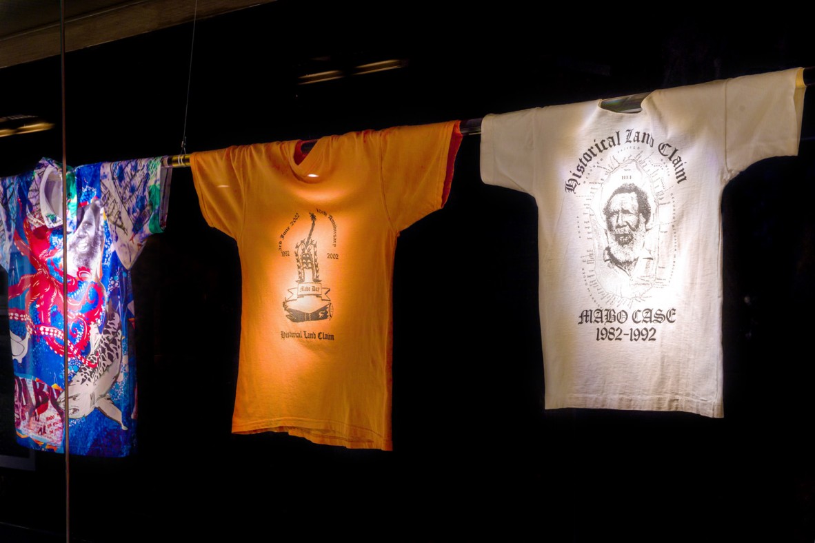 Three shirts with prints celebrating the Mabo High Court case hanging by their sleeves on a pole in a glass case.