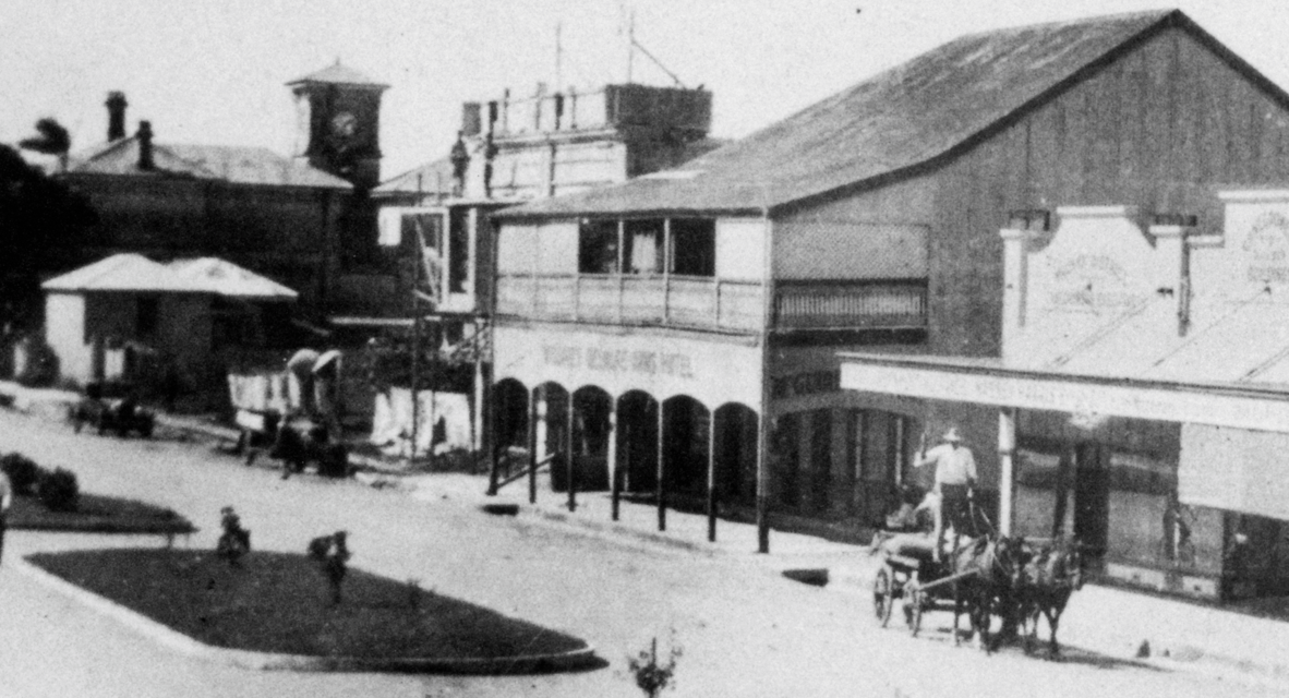 Black-and-white historical street scene showing a small town centre with early shopfronts and verandahs. A horse-drawn carriage with a driver stands in the road, while a few pedestrians are scattered nearby.