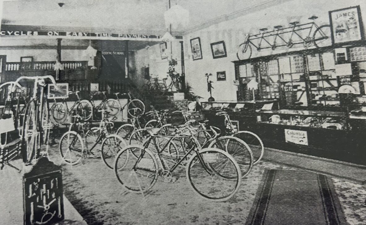 Black-and-white photograph of a late 19th-century bicycle showroom filled with upright bicycles arranged across the floor. Glass display cabinets line the walls, holding parts and accessories, while framed items and signage hang above. A multi-seat tandem bicycle is mounted high along the wall.