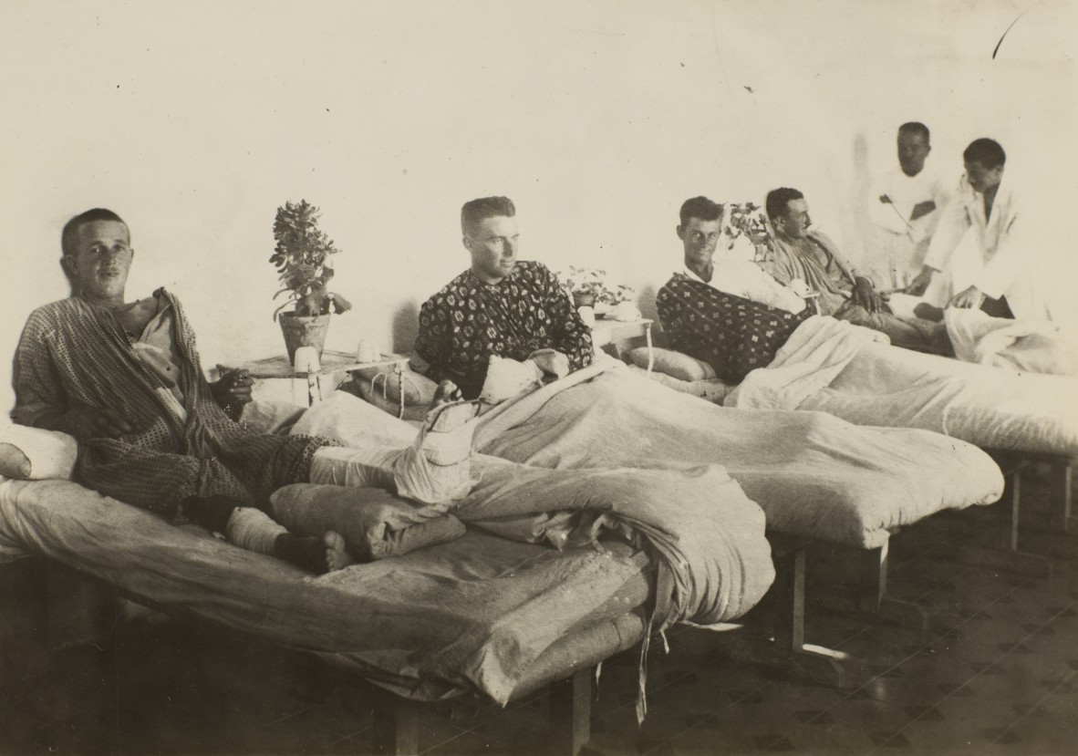 Four men in a hospital ward, in beds lined up against a wall, being attended by two men in white coats. The man in the foreground has his left leg in a splint.