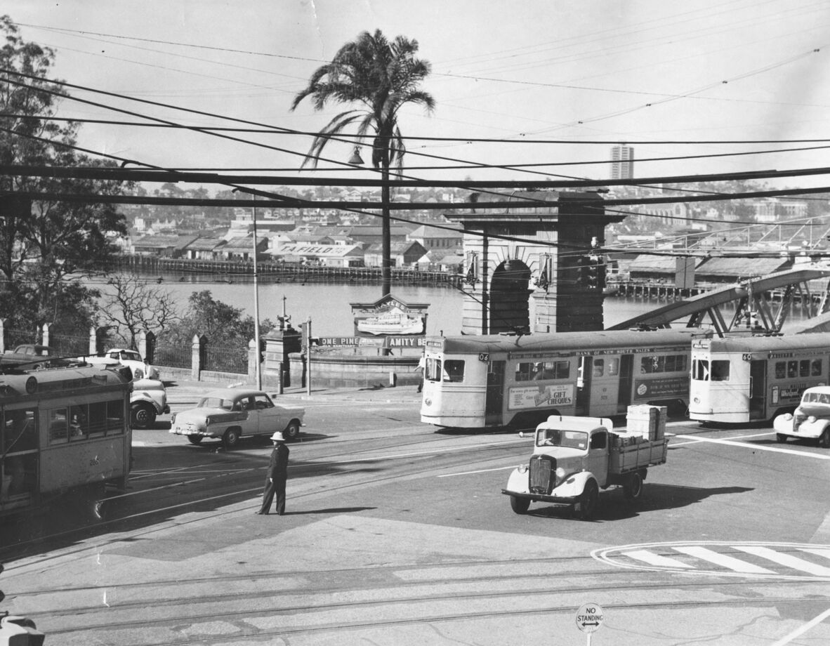 Black-and-white photograph of a busy street scene at the northern end of Victoria Bridge, Brisbane, circa 1960. Trams, cars, and a small truck converge at an intersection while a uniformed traffic officer stands in the roadway directing vehicles.