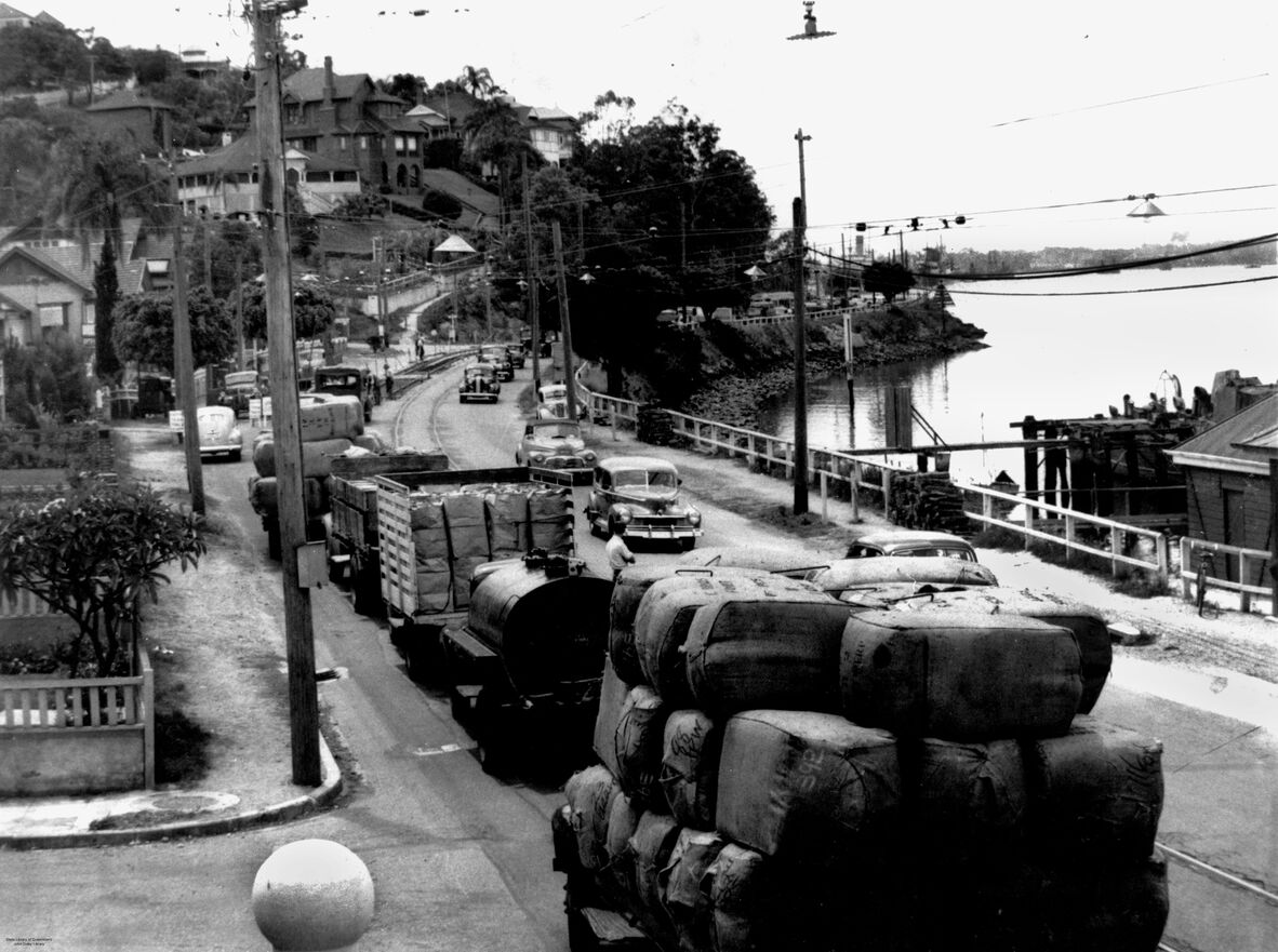 Black-and-white photograph of heavy traffic along a riverside road in Brisbane, 1949. Trucks loaded with goods, cars, and tankers queue along a narrow roadway beside the river, with hillside houses, power poles, and overhead lines framing the busy, industrial scene.