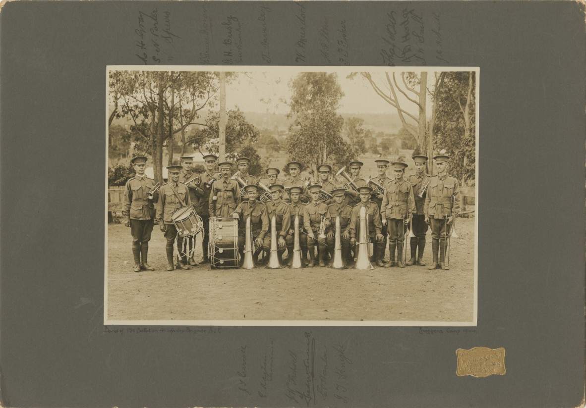 Group photo of twenty soldiers in uniform, holding brass instruments and drums, trees in background, dirt ground cover. 