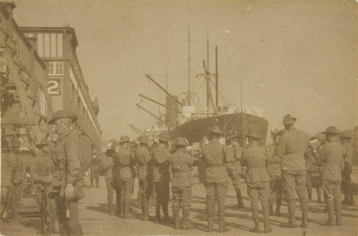Faded picture of a group of men in uniform facing away from the camera toward a large ship, with buildings to the left.