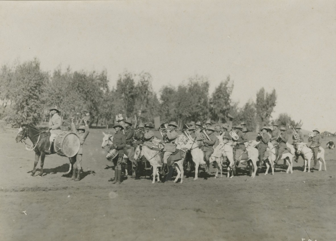 Group of approximately 20 soldiers in uniform, facing left of camera point of view, riding donkeys. Soldiers playing brass instruments and drums. 