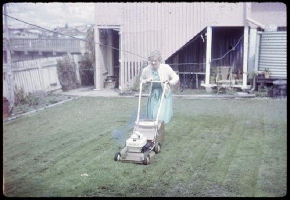 Woman mowing the lawn with an electric lawnmower. In the background is a water tank and the suburban skyline. 