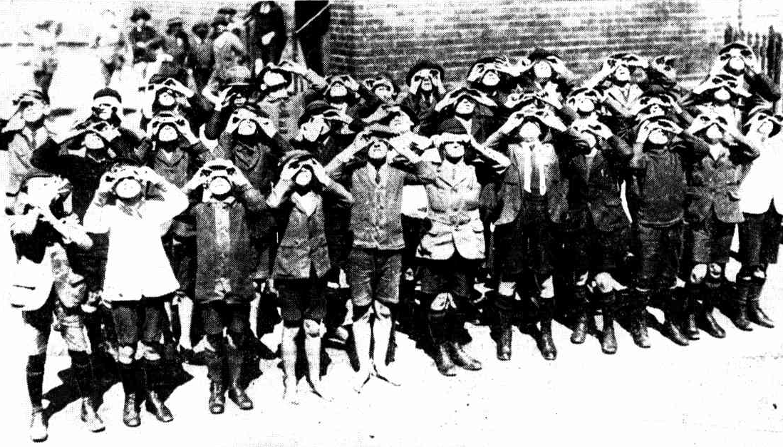 A group of children stands closely together, all wearing protective eyewear and looking up towards the sky, possibly viewing a solar event. The setting is outdoors, with a brick wall in the background.