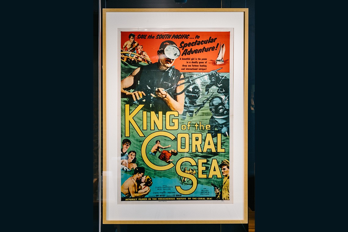 Colour lithograph poster promoting the 1954 Australian film King of the Coral Sea which was shot on location on Thursday island and in the waters off Green Island near Cairns. The coloured poster features illustrations of a sailing boat, deep sea divers wearing helmets, a man in a scuba mask holding a spear gun and a man and woman in swimsuits kissing passionately.