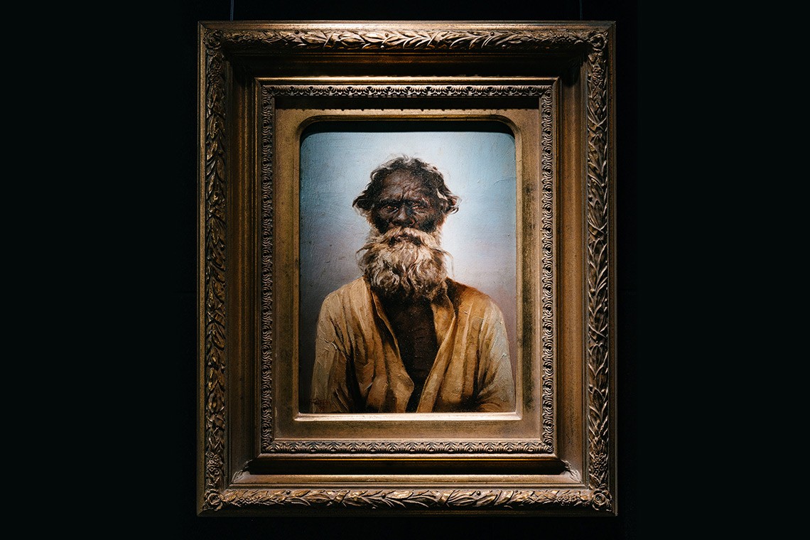 This oil painting by Swedish born artist, Oscar Fristrom depicts Kirwallie or "King Sandy", a well-known Aboriginal elder of the Brisbane region.  The painting is housed in an ornate gold coloured frame.  it is a head an upper body depiction of an elderly Aboriginal man with a full grey white beard.  He is wearing a white shirt which is open to his chest which shows signs of tribal scarification. 