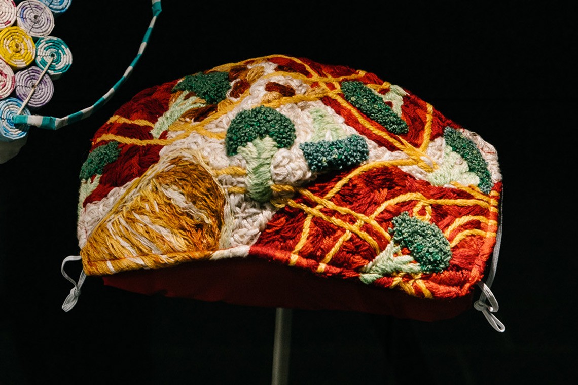 The Covid 19 mask “Covid-19 made me buy it” reflects on supermarket shelves being stripped bare during lockdowns. The face maks is constructed from cotton embroidery floss on calico with a milled linen backing and an elastic strap. It features green heads of broccoli on a red and cream background.