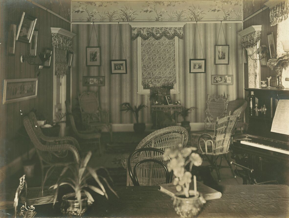 A vintage photograph of a decorated parlour with striped wallpaper, hanging framed pictures, wicker chairs, indoor plants, and a piano on the right.