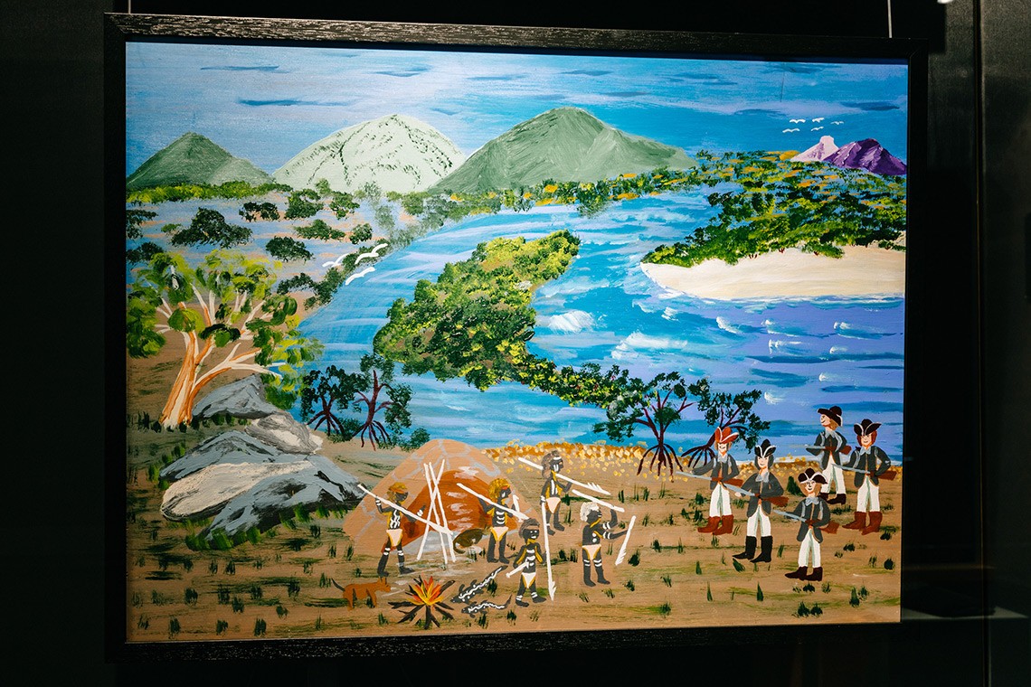 The painting, "Defending Our Camp" depicts the First Nations’ oral history of Captain Cook’s landing at Endeavour River. The acrylic on plywood painting depicts the landscape of the Endeavour River with the green mountains in the background and the blue river winding through the land. In the foreground a group of Bama people with spears are standing in front of their humpy and are facing Cook's men who are carrying guns.