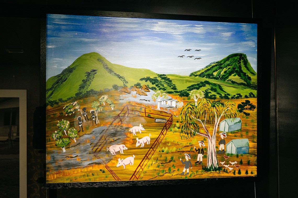 The painting, ""Burning the Pigs"" depicts the First Nations’ oral history of Captain Cook’s landing at Endeavour River. The painting is acrylic on plywood and depicts the green hills of the endeavour river in the background. The foreground features a burned pig pen containing four pigs, with Bama people on the left and Captain Cook's men on the right with their tents and chickens.