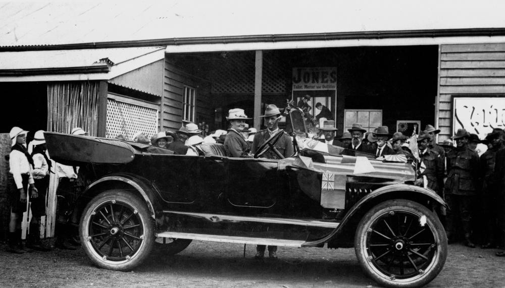 Image of Mrs Wheeler sitting in car in front of buildings with a group of children and adults spread out behind the car, ca.1919