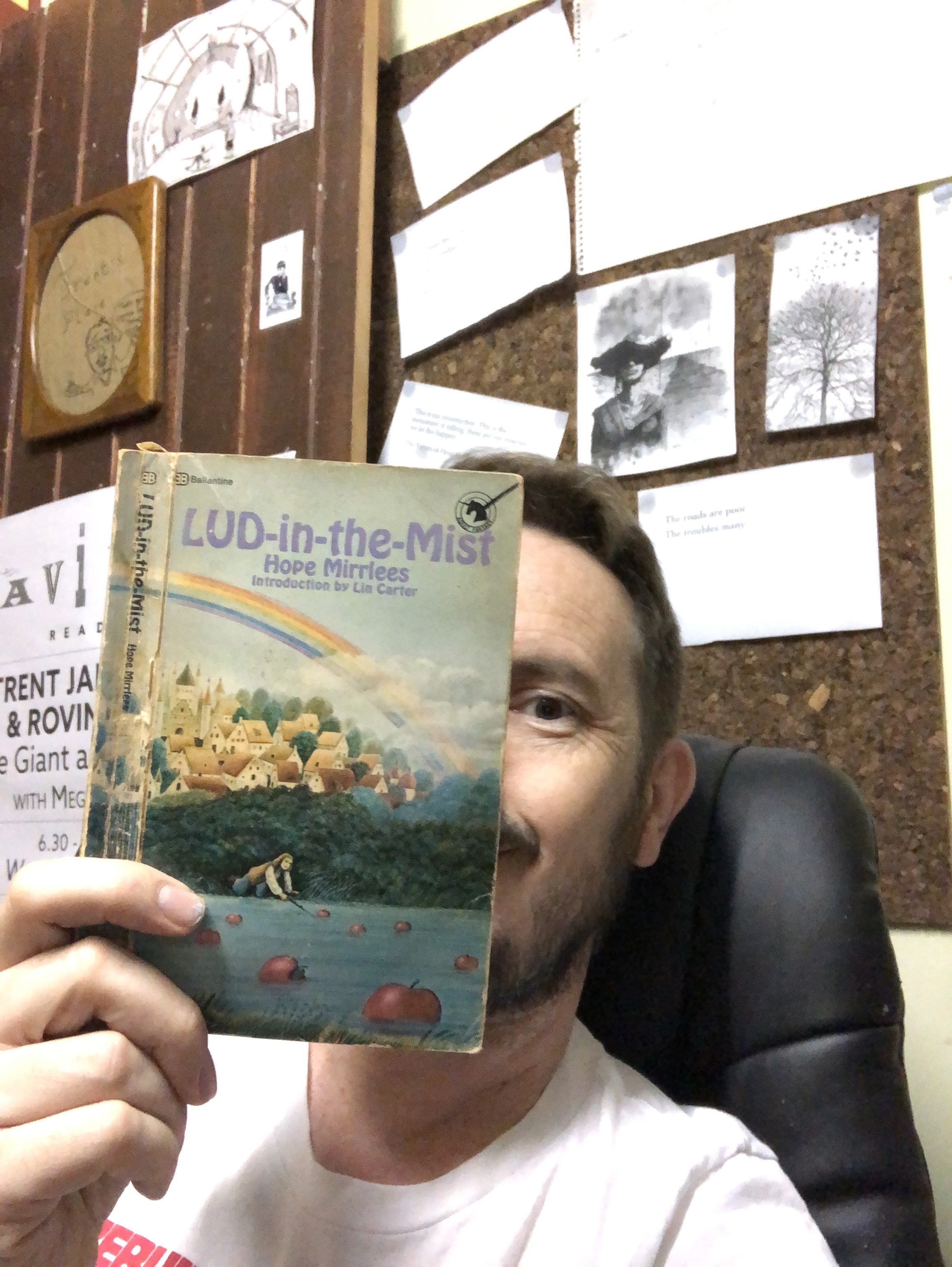 Trent Jamieson holds up a copy of Lud-in-the-mist by Hope Mirrlees. Trent sits in front of a pin board