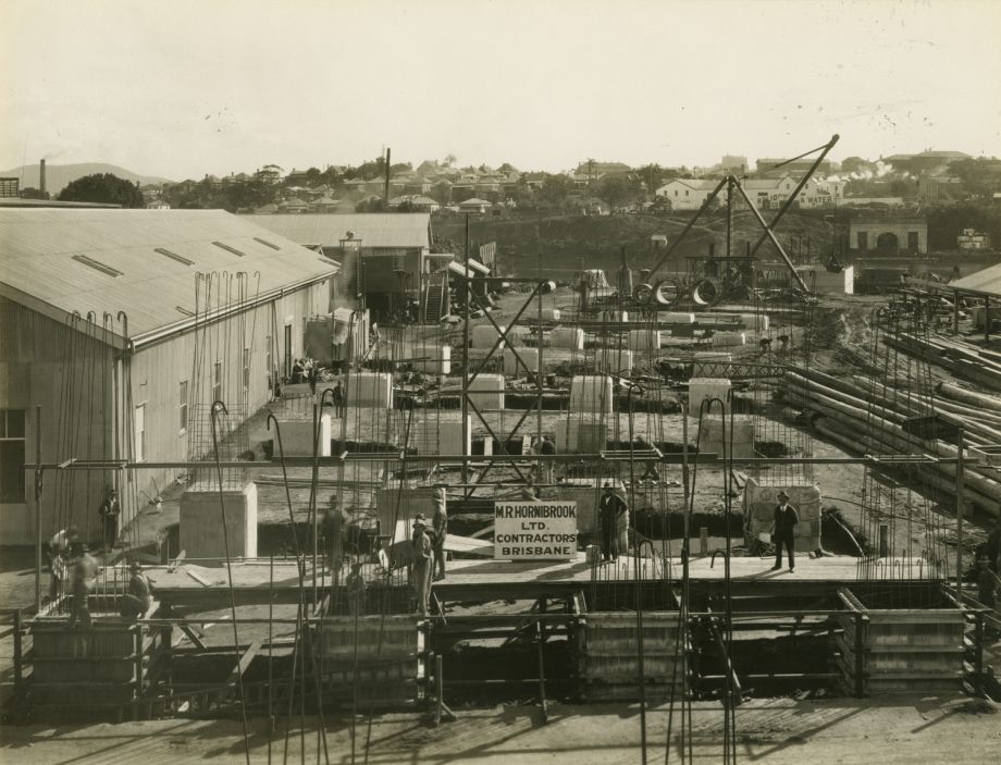 Concrete work in progress to become arches supporting the road of the Bridge on the south side. Across the Brisbane River the Helidon Spa company warehouse and housing along Coronation Drive can be seen. Hornibrook's site office is by the river in the middle distance at left, with construction workshops in the left foreground.