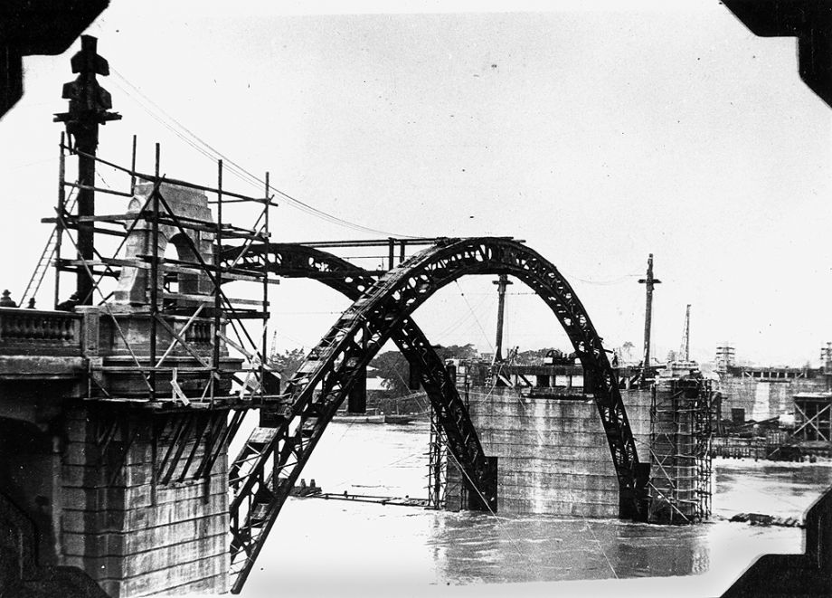 Construction of the William Jolly Bridge, then called Grey Street Bridge, under construction as the Brisbane River flooded in 1931, photographed 7 February 1931. Each rib of the three spans consisted of structural steel fabricated under a sub-contract by M. R. Hornibrook Ltd. to Evans, Deakin and Co., Brisbane. This steelwork was designed to support the concrete during construction without falsework.