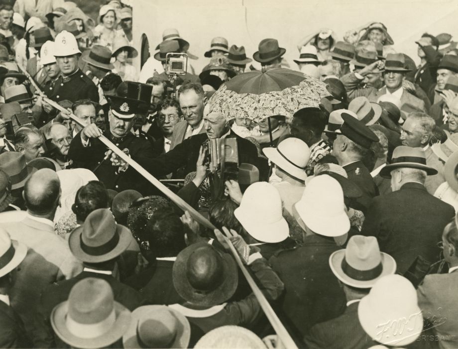 The image captures the moment of cutting the blue ribbon to open the Grey Street Bridge. The golden scissors had been handed to him by Manuel Hornibrook, standing behind him, not wearing a hat. An umbrella is held over the Lord Mayor W.J. Greene and the Lady Mayoress next to him. Local police and military police assist with managing the crowds.