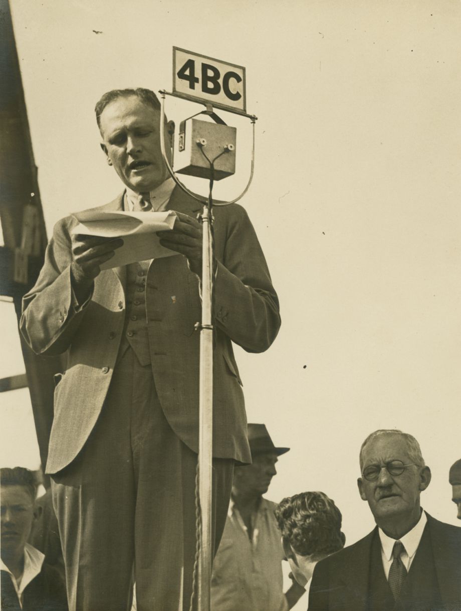 Manuel Hornibrook, owner of the company that built the William Jolly Bridge, stands behind a large boxy 1930s microphone with speech notes in hand.