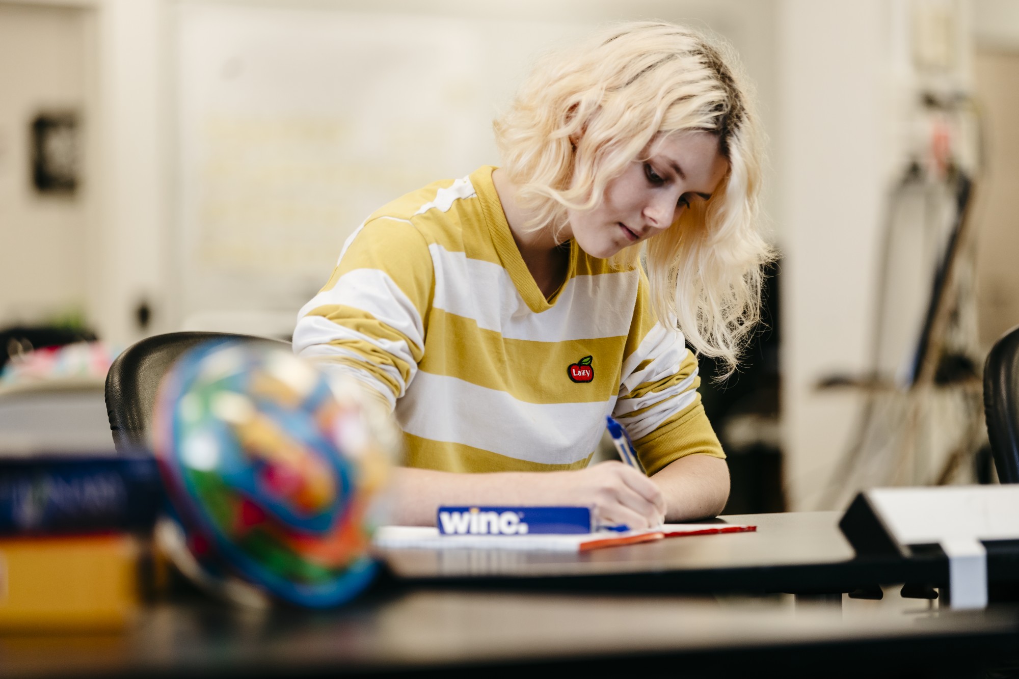 Person wearing a yellow striped shirt writing on paper while sitting at a desk.